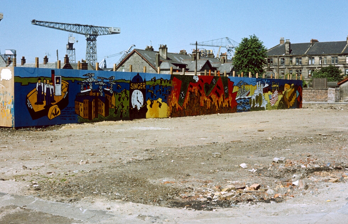 Image of colourful artwork of Clydebank imagery on the wooden fencing on the outskirts of a shipyard, with crane visible in distance.