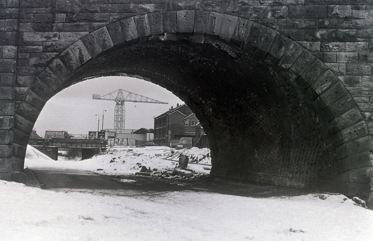 B+W Image of a brick arch-way bridge from underneath, with a crane visible through the arch.
