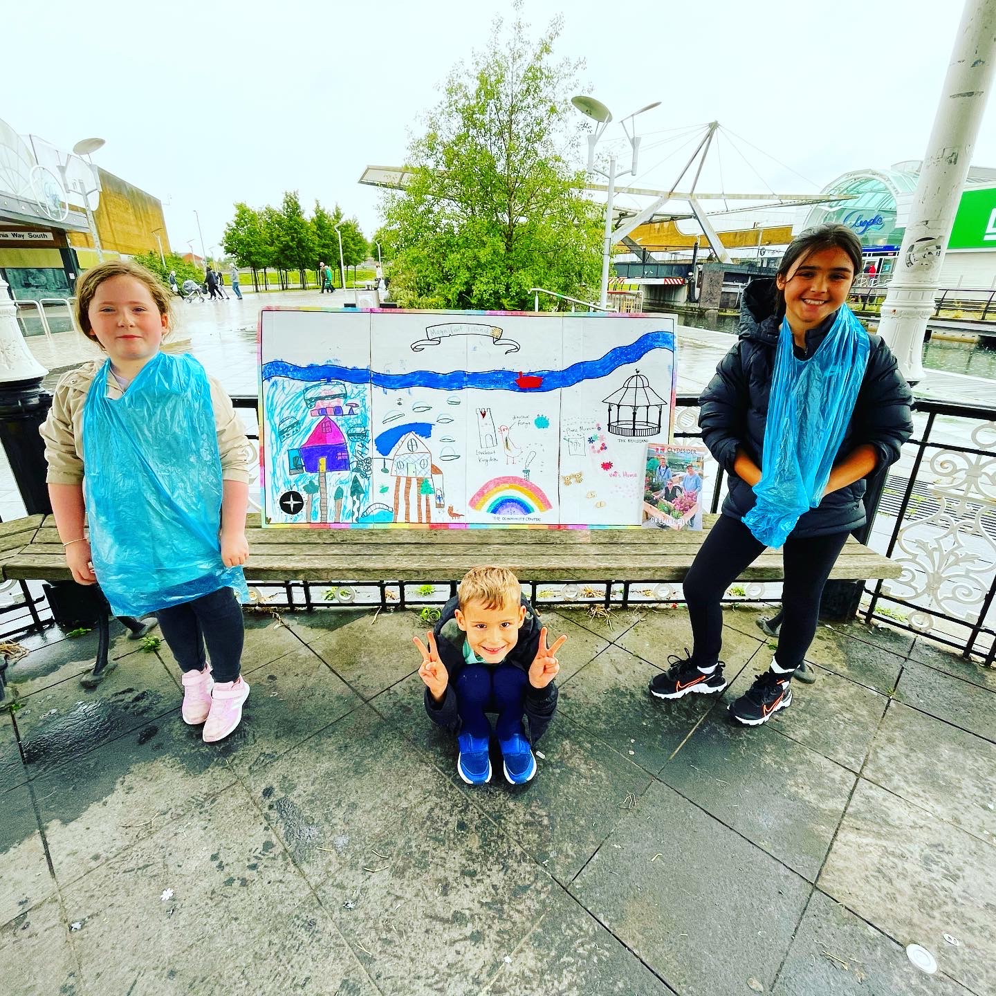 Susan Laws 3 children standing in the bandstand in the heart of Clydebank Shopping Centre, smiling next to a colourful artwork.