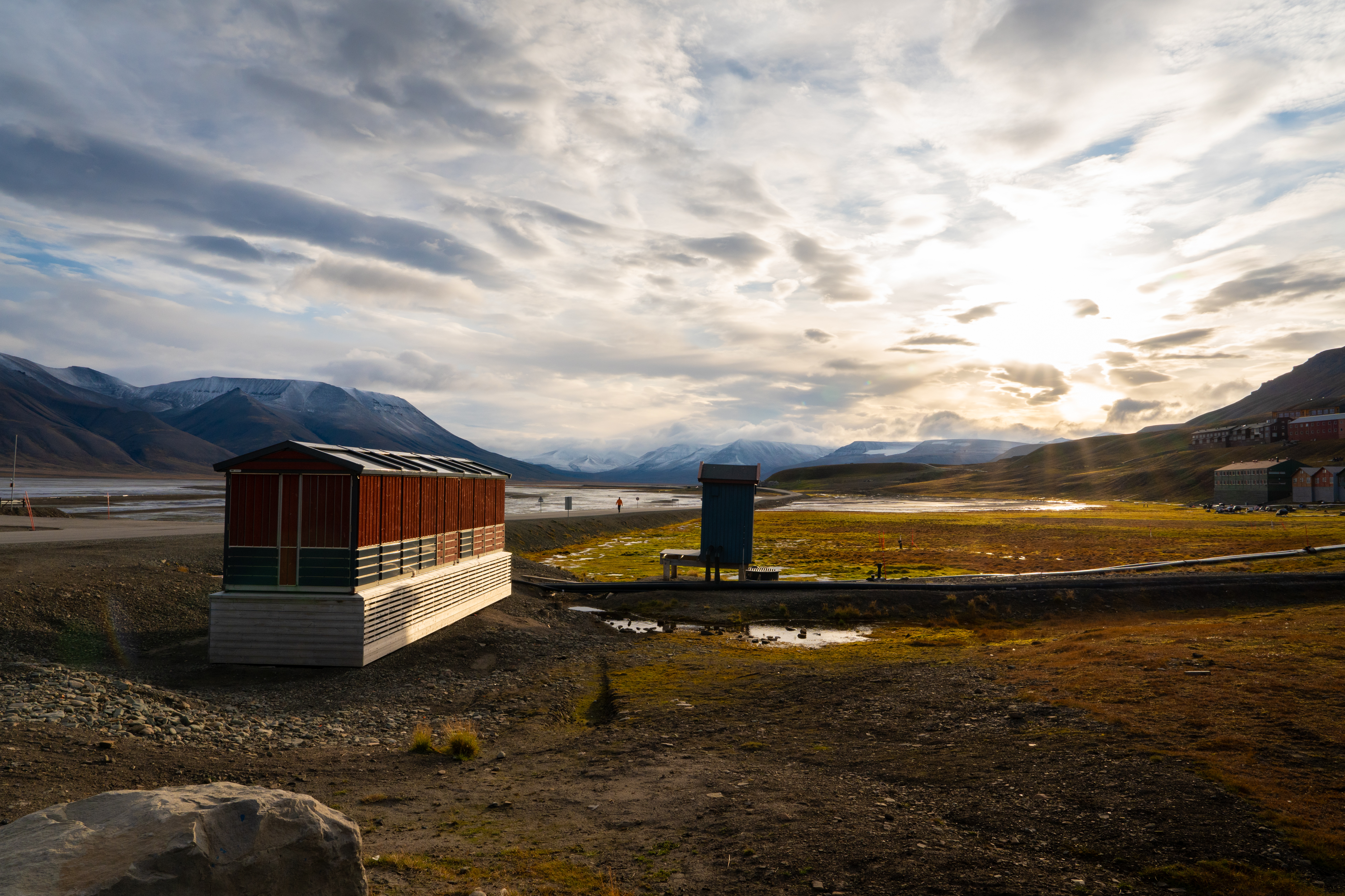 landscape photo with wooden building to the fore and hills in the distance. Sun is shining through the clouds in the sky