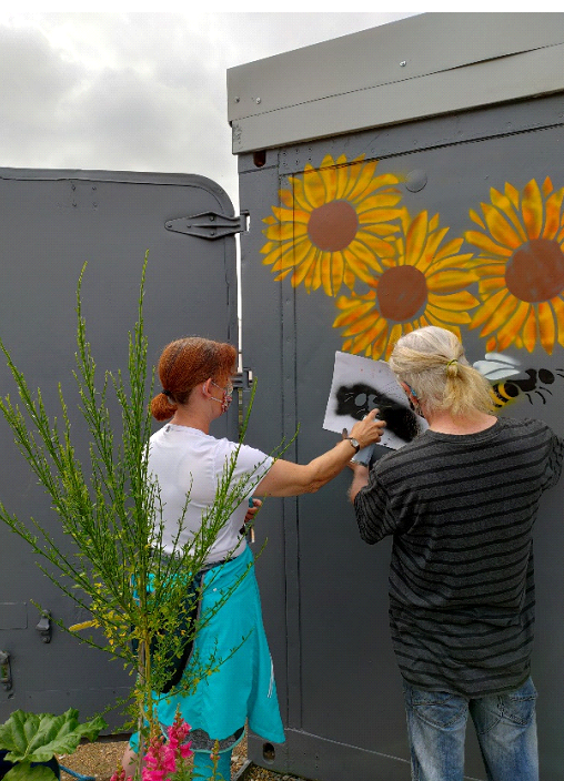 A lady and man faced away from camera, painting sunflower designs onto side of an outside container.