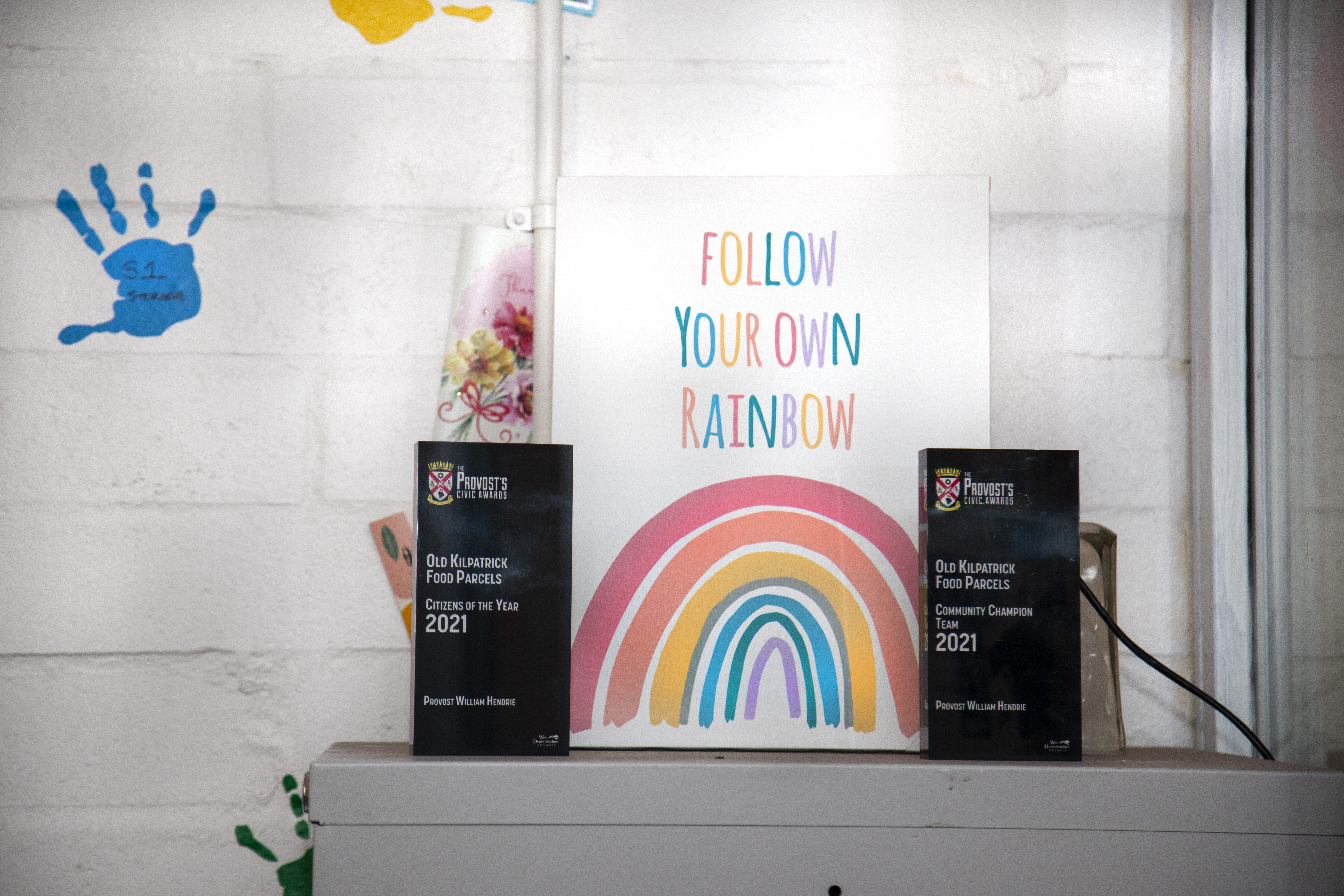 A shelf with a white wall background, ontop is 2 awards titled towards OKFP and a poster featuring a rainbow and the quote "Follow Your Own Rainbow"