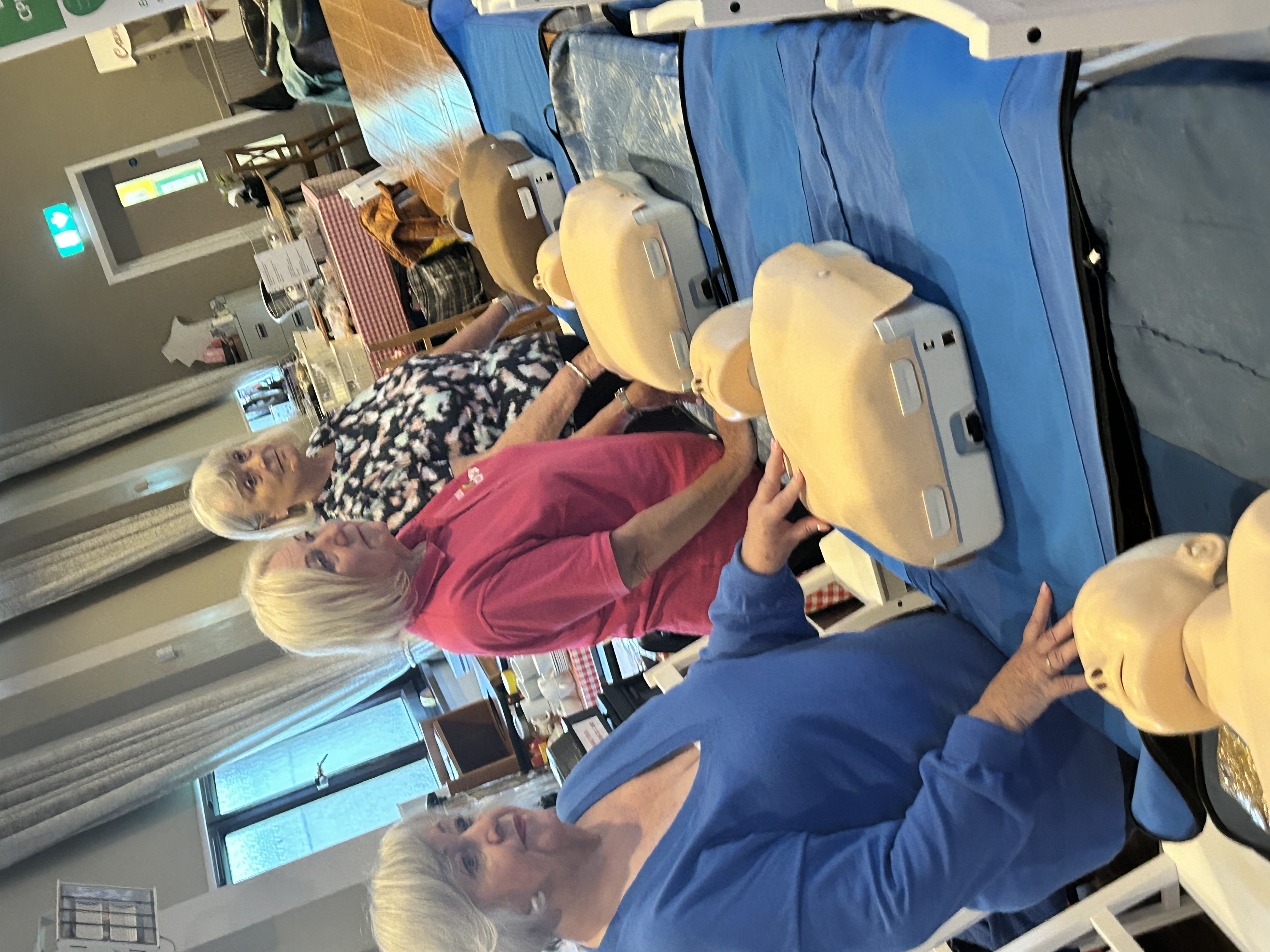 Three women standing next to tables with CPR dummies in front of them