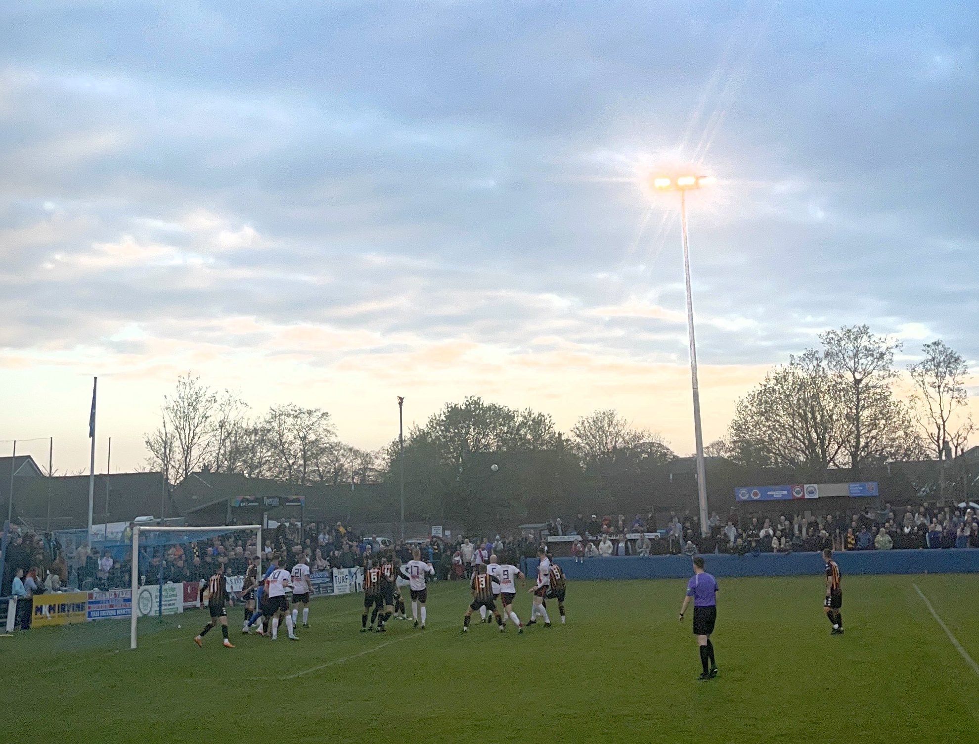 footballers gathered together on a pitch waiting to see if the ball in flight will make it to the goal