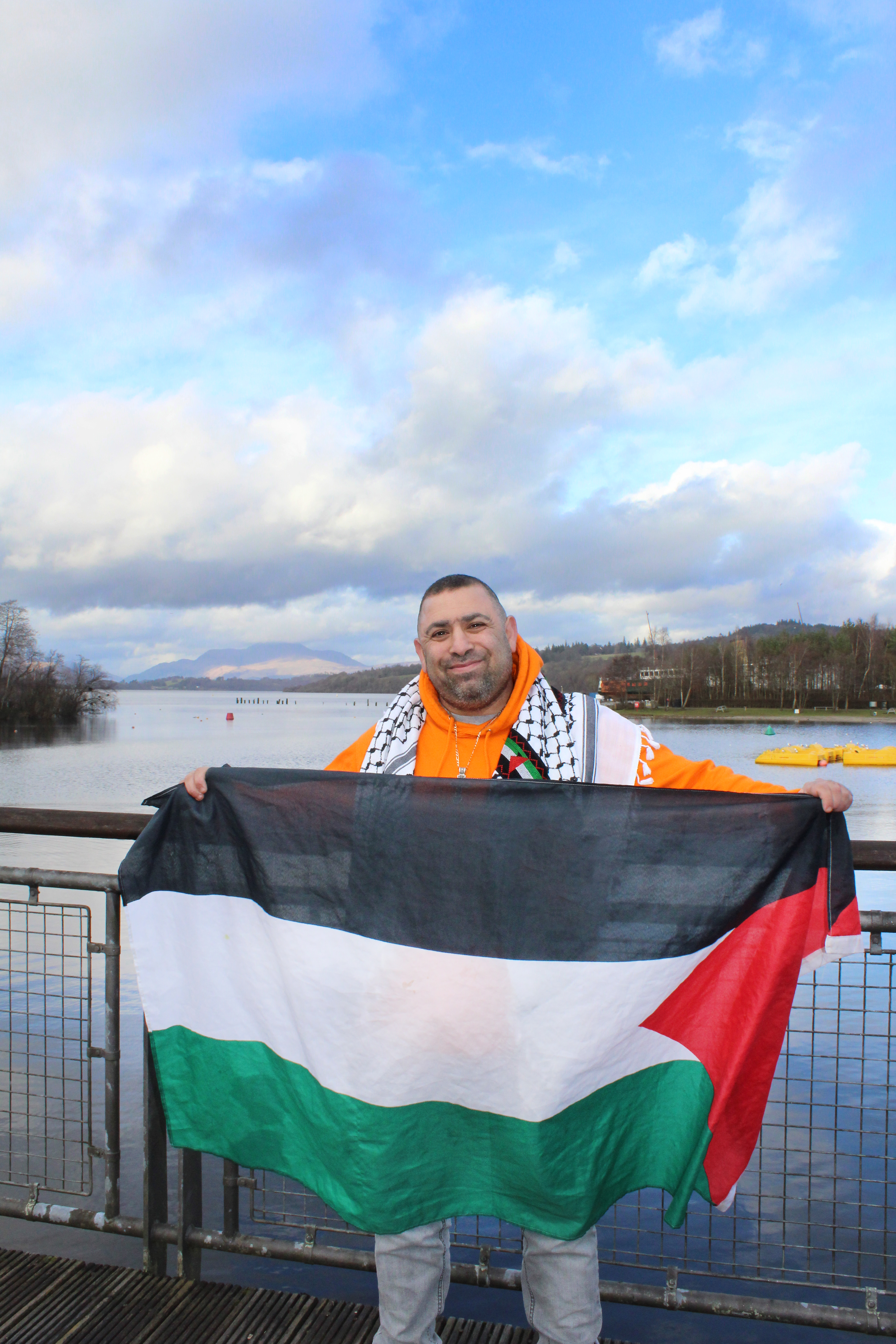 Simon standing holding Palestinian flag with Loch Lomond n background