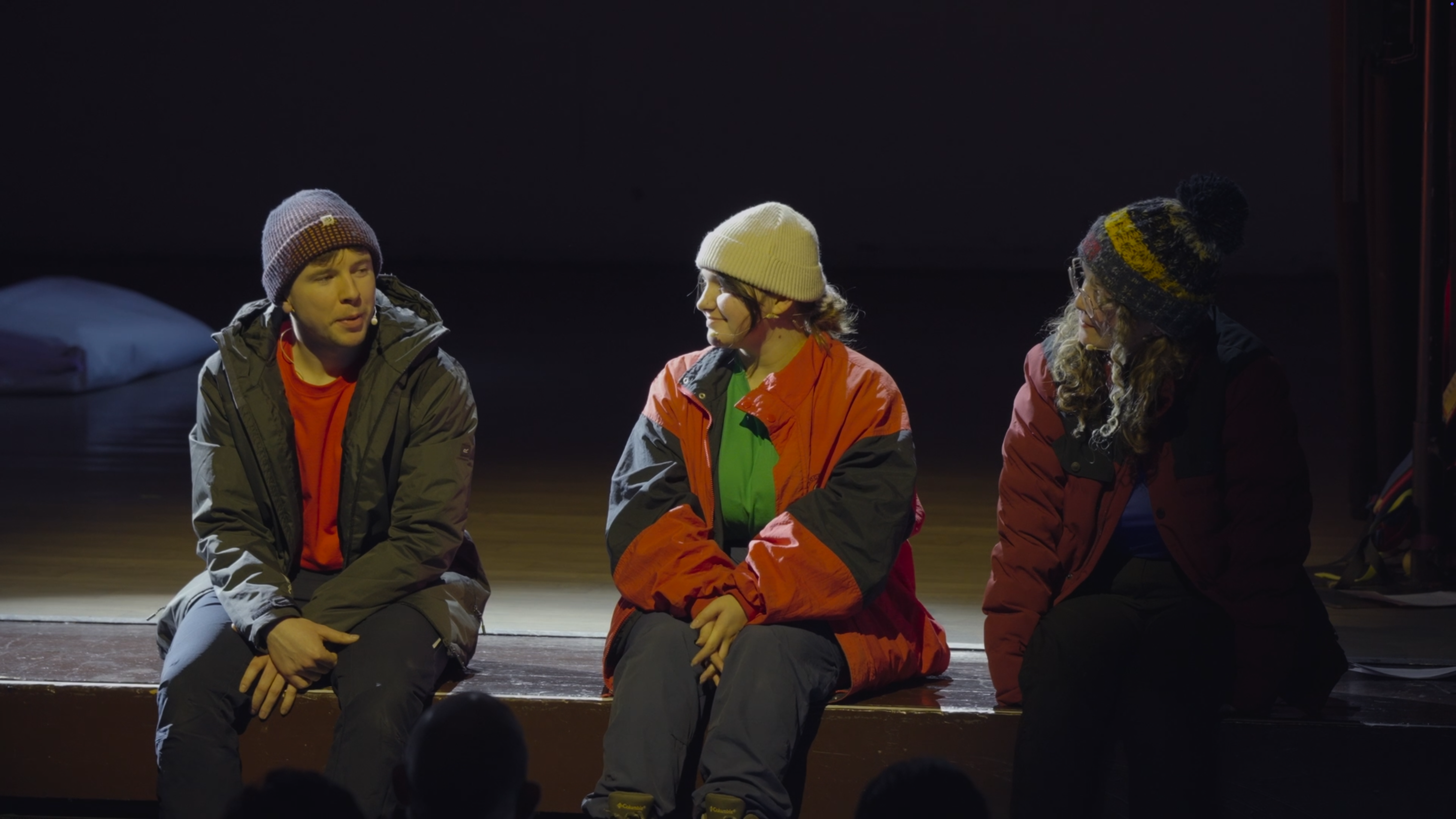 Three actors sitting on the edge of a stage, talking to one another during their performance
