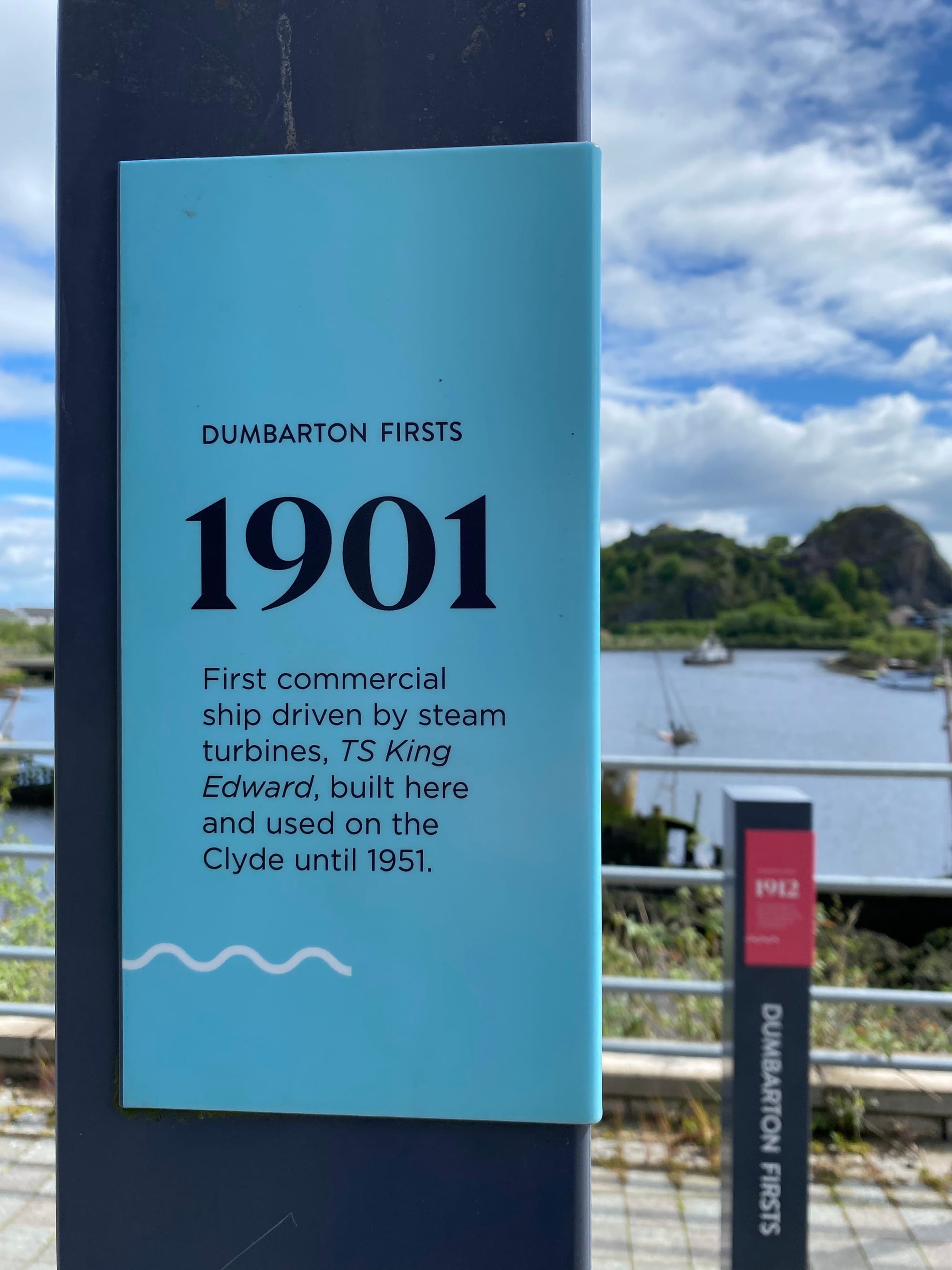 A sign saying 'Dumbarton Firsts 1901 - First commercial ship driven by steam turbines, TS King Edward, built here and used on the Clyde until 1951' with Dumbarton castle in the background