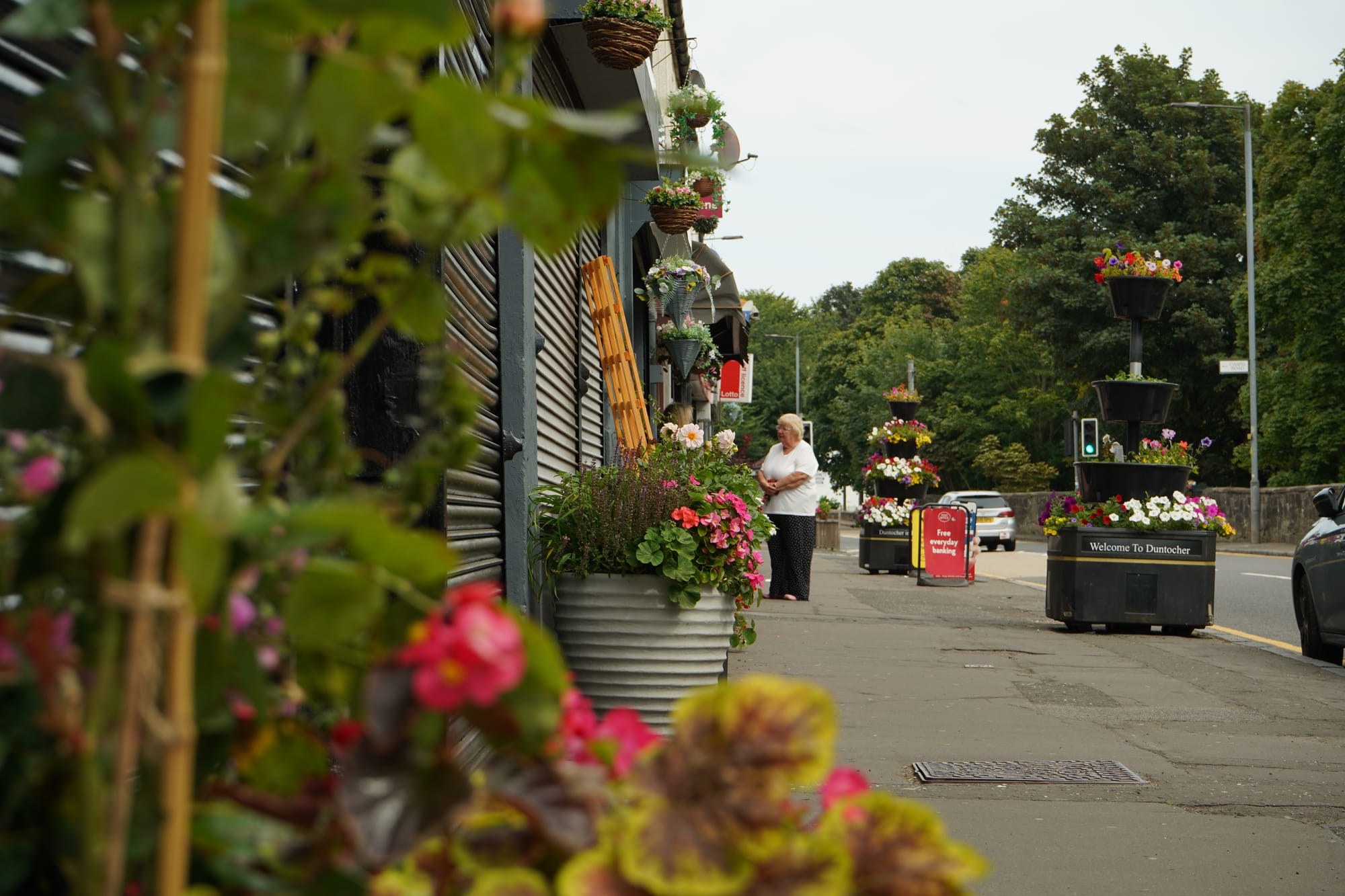a small section of the main street in Duntocher with lots of planters in bloom visible